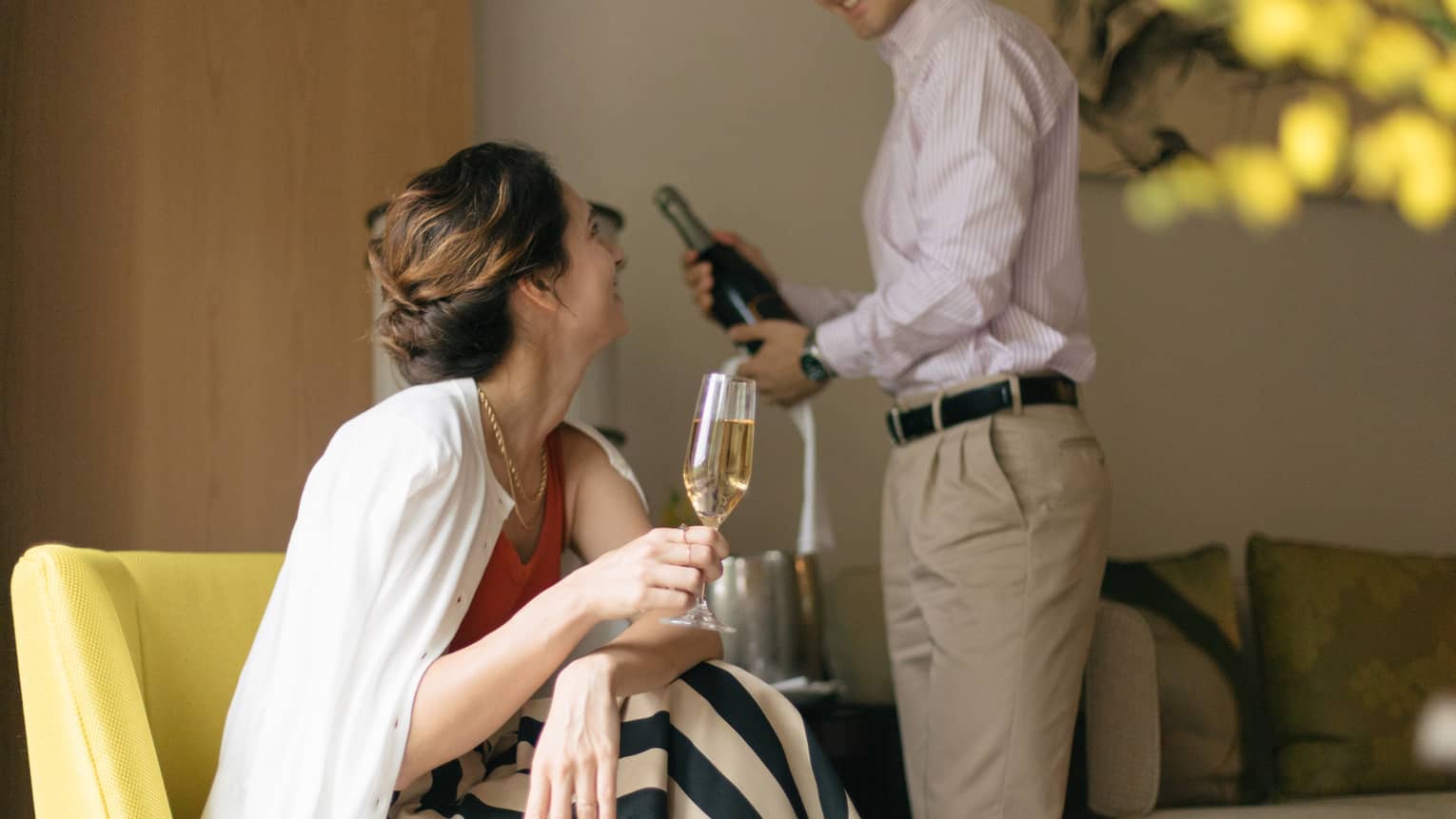 Person smiling and holding a glass of champagne while another person opens a bottle