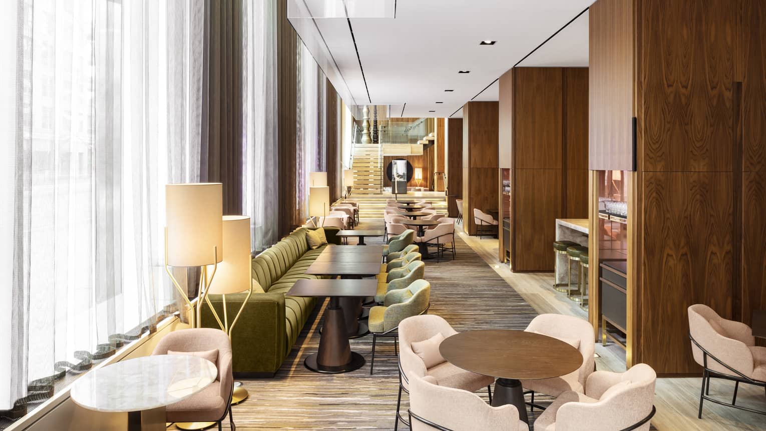 A long room lined with upholstered sofas and chairs, wood tables and windows