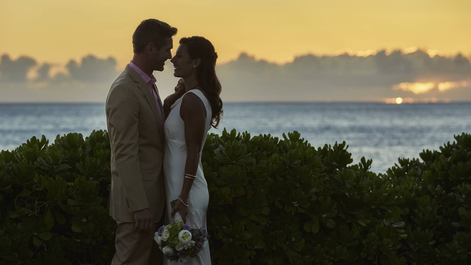 Silhouettes of bride and groom leaning in for kiss against sunset