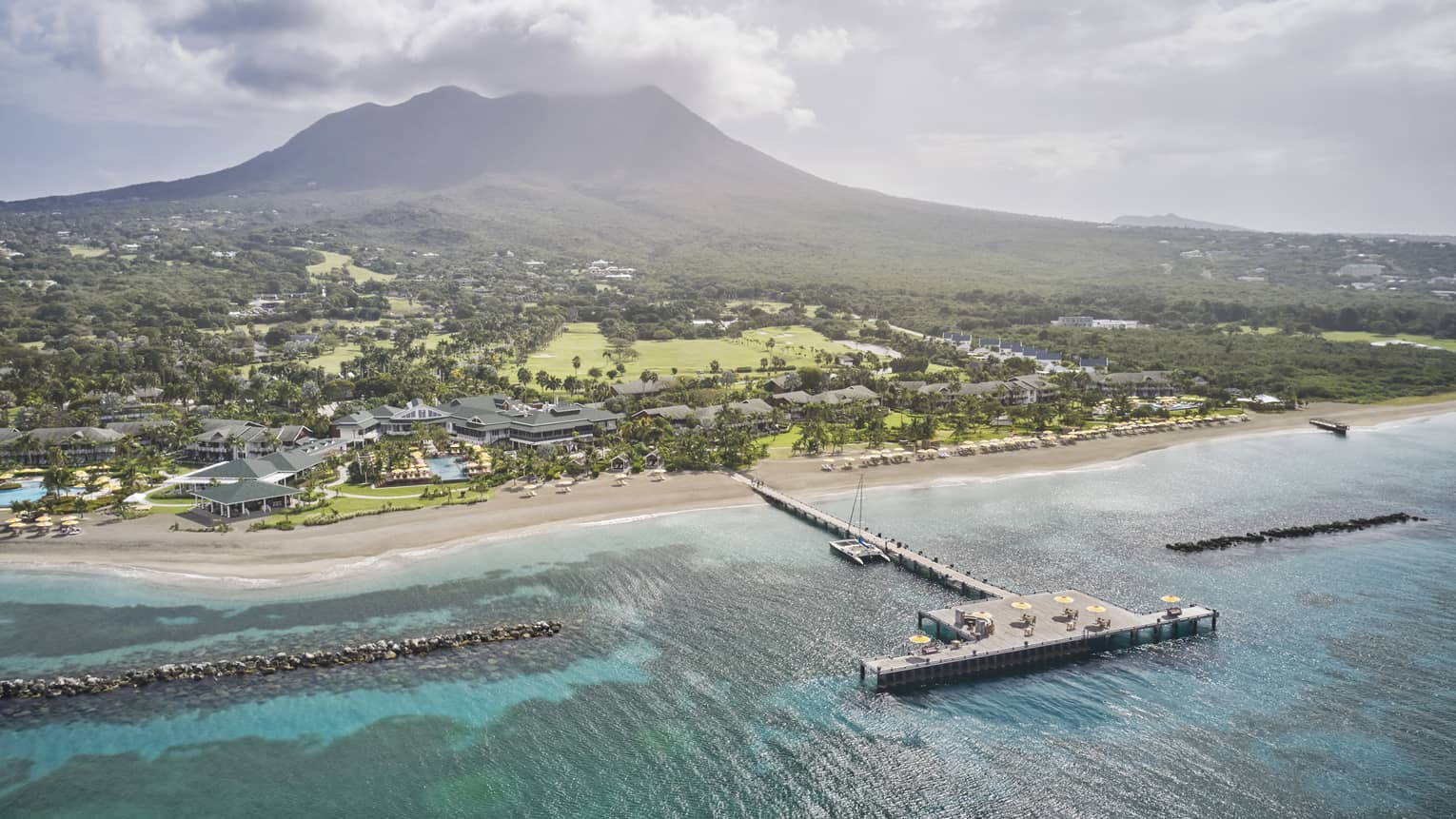 Aerial view of a resort property and dock on a beach shore with a mountain in the distance.