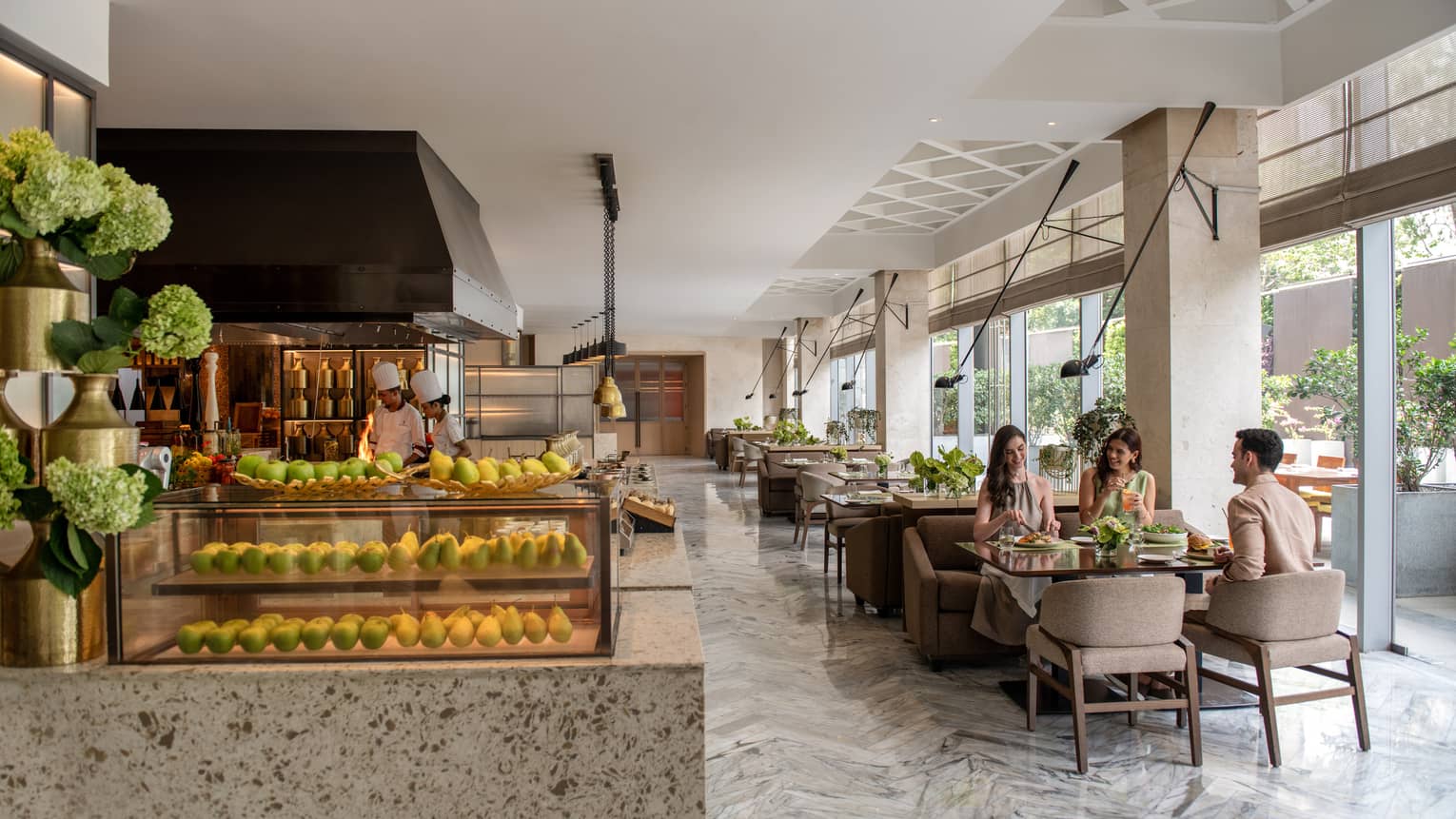 Three people dine at restaurant with marble floors and natural light, next to open show kitchen