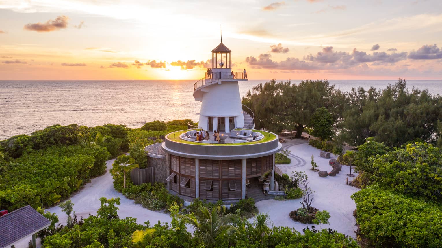 Lighthouse nestled among greenery with people on different levels, overlooking the ocean at sunset