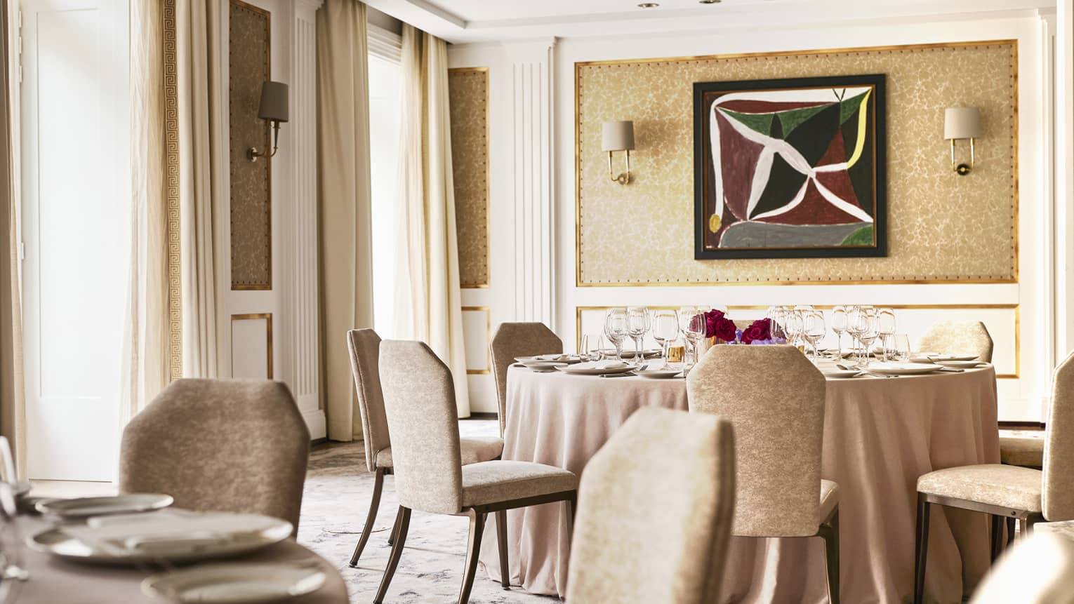 A room filled with natural light is decorated in light, warm, neutral colors and has large windows, wall lighting, tables and chairs, and a piece of art on the wall