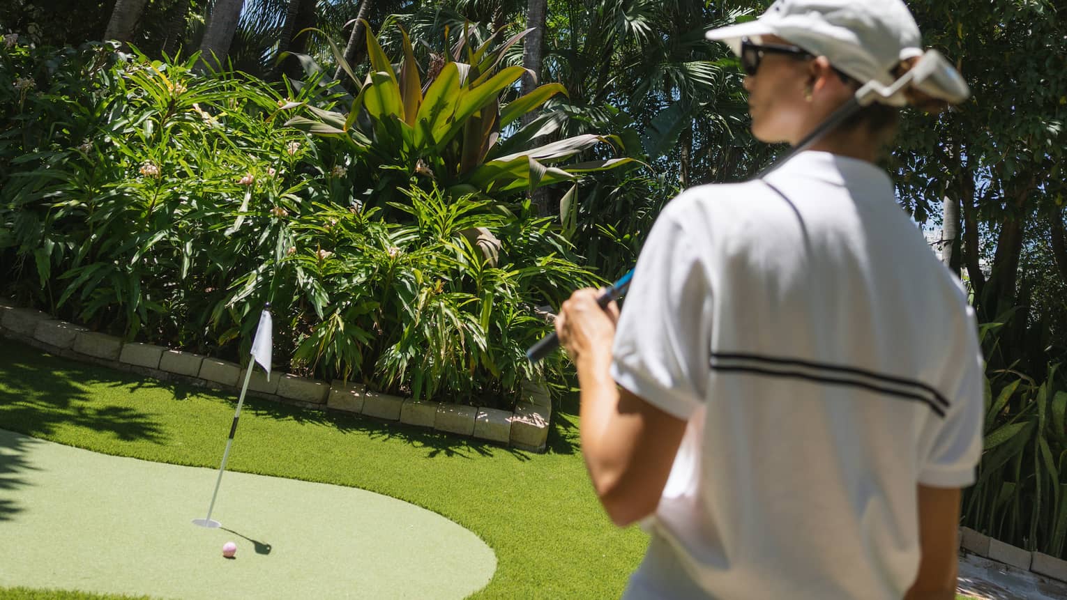 Golfer wearing white sweater vest, white skirt and white hat walks toward a putting green surrounded by palm trees