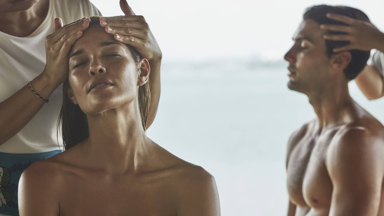 Close-up of woman and man with eyes closed as masseuses massage their temples