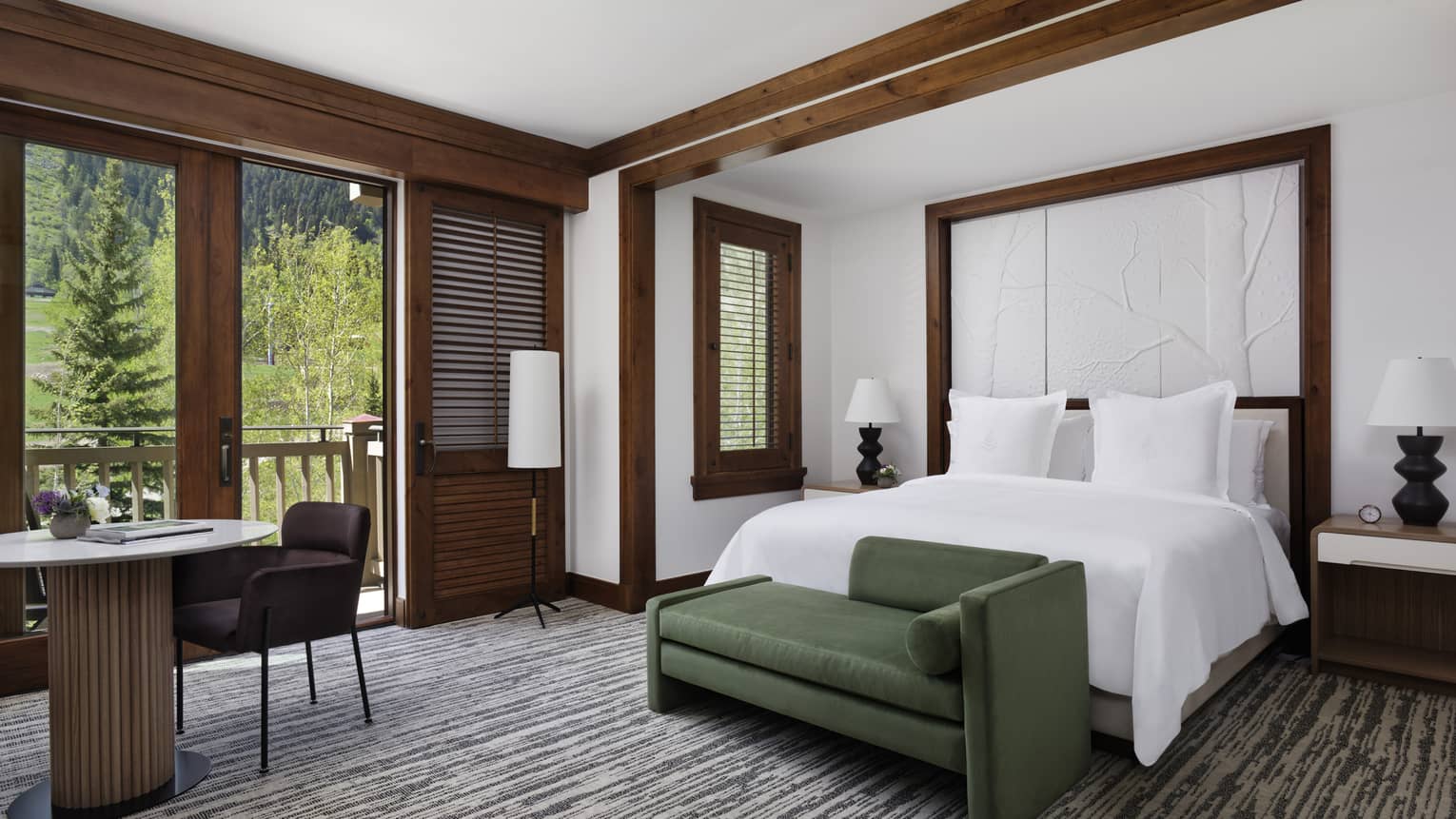 Hotel room with king bed, green chaise, table and two chairs, and balcony view of forest