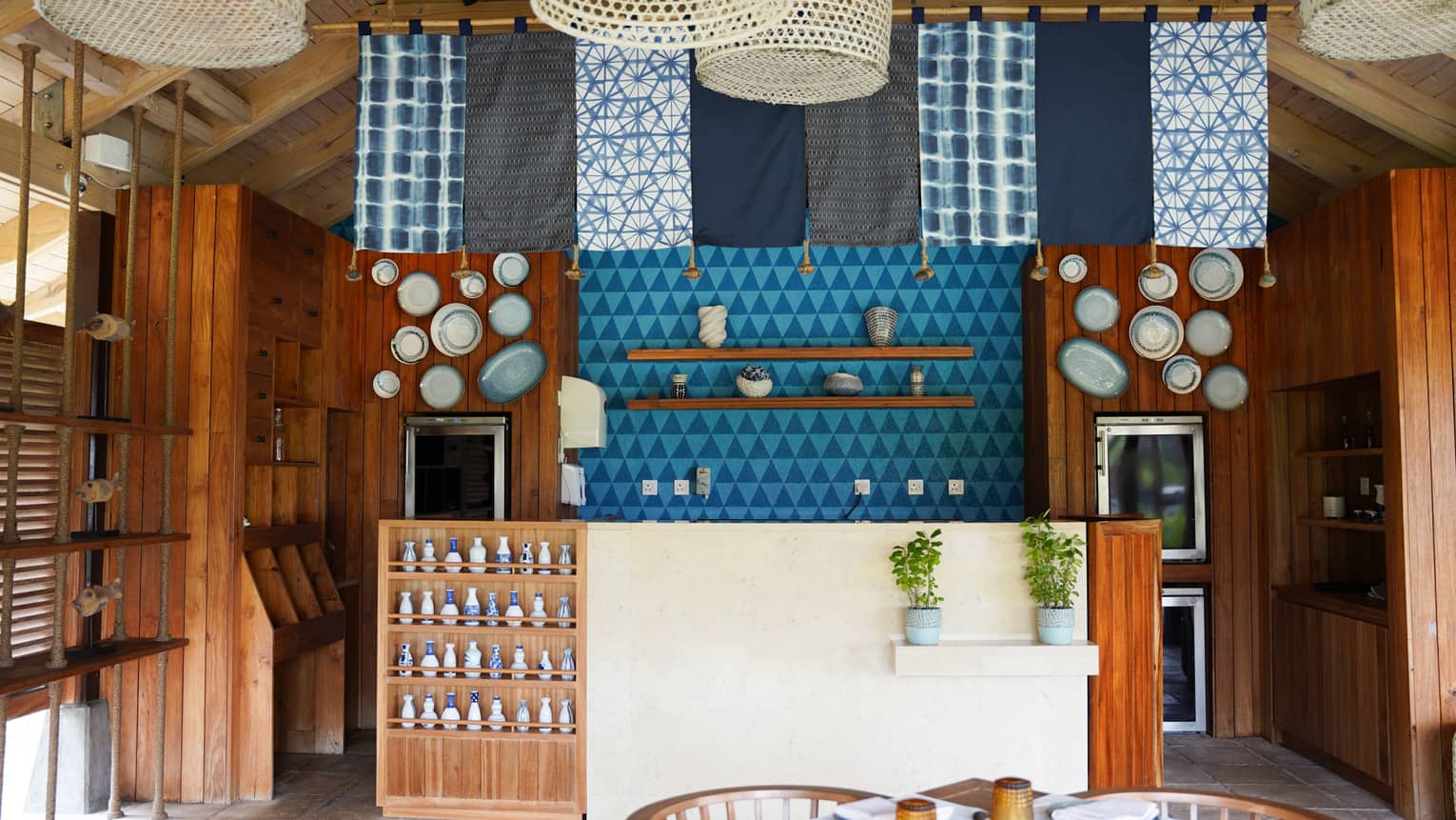 Casual restaurant with an island vibe, with blue and white fabric panels and rattan lights hanging from ceiling over teak and stucco bar