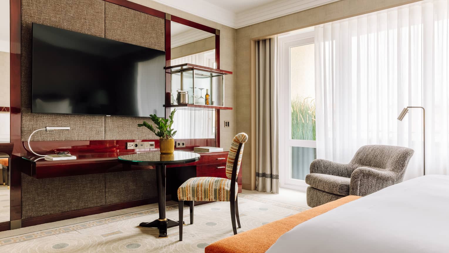 A modern hotel room with a large bed, a flat-screen TV, a work desk, and a sitting area with chairs and a small table. The room features large windows with city views.