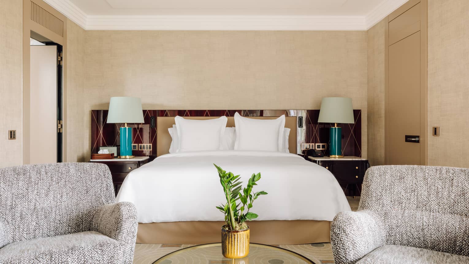 Hotel bedroom with a large bed, two armchairs, a round glass coffee table and bedside tables with green lamps.