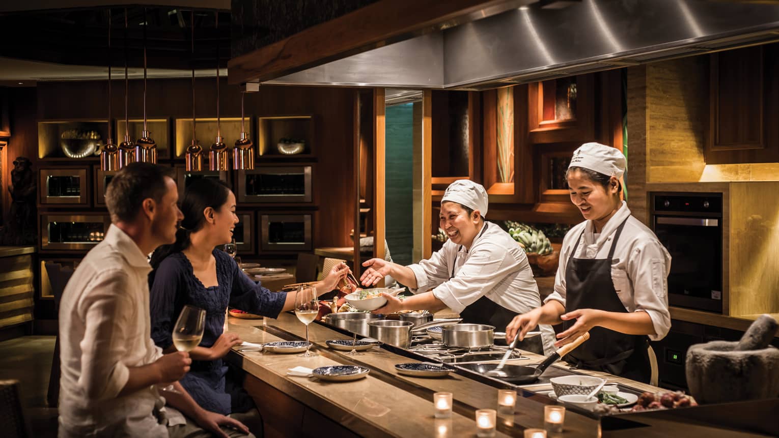 Two chefs smile and laugh with a couple as they prepare them private dinner