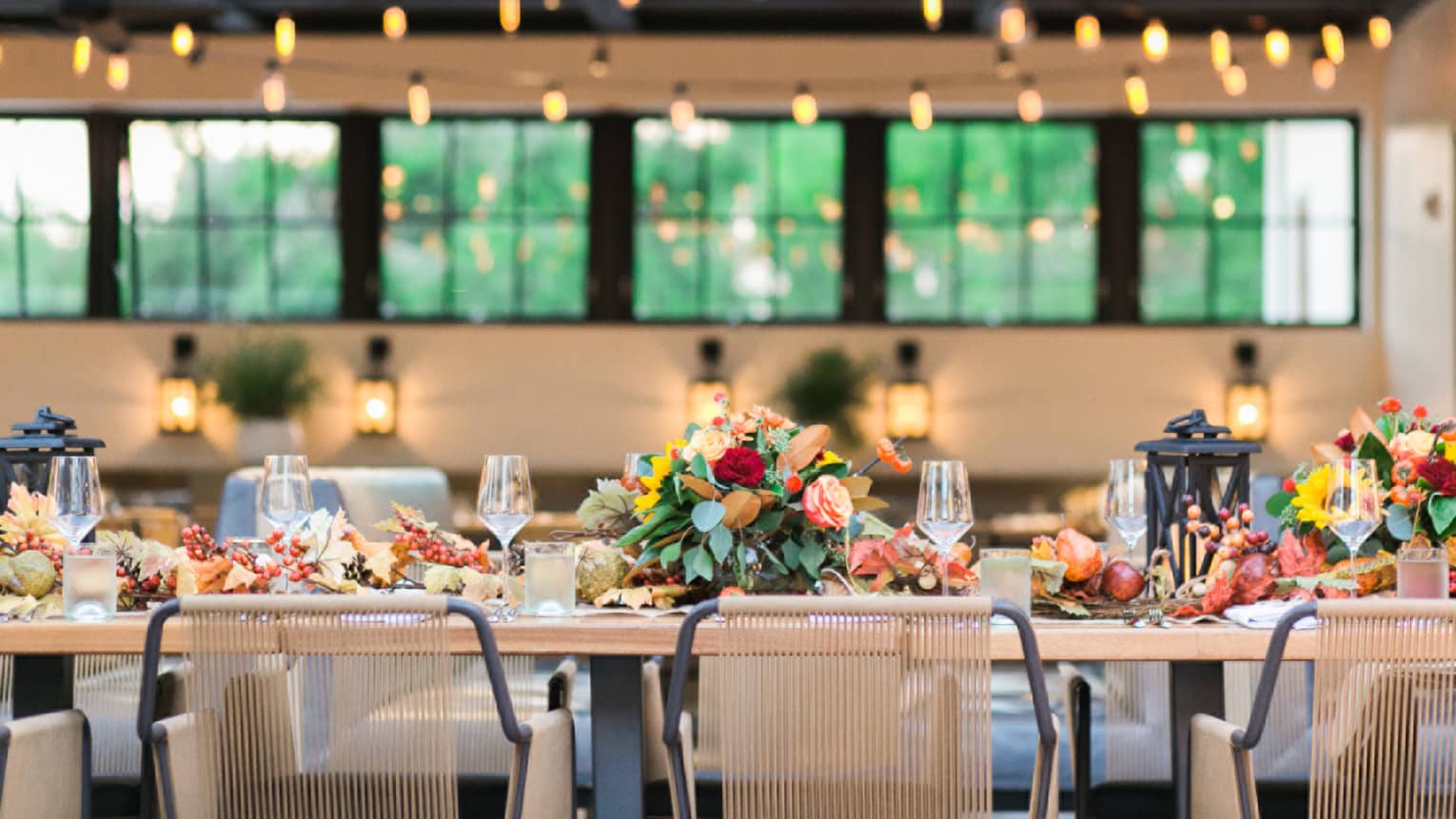 Long outdoor dining table with harvest flower arrangements under strings of bulb lights during day