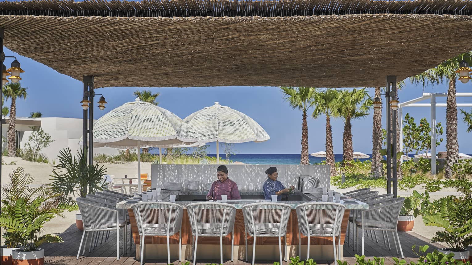 Two staff members behind beach bar with thatched roof, white rattan chairs, water view