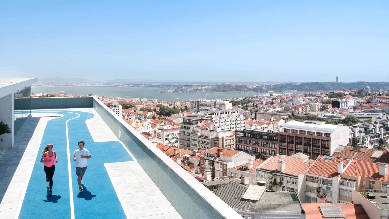 Two people run on blue, rooftop running track, overlooking Lisbon
