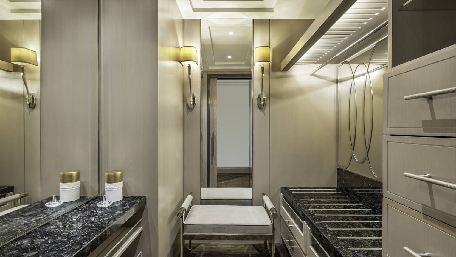 Elegant walk-in closet with built-in drawers, black granite countertops, soft lighting and a cushioned bench under a large mirror