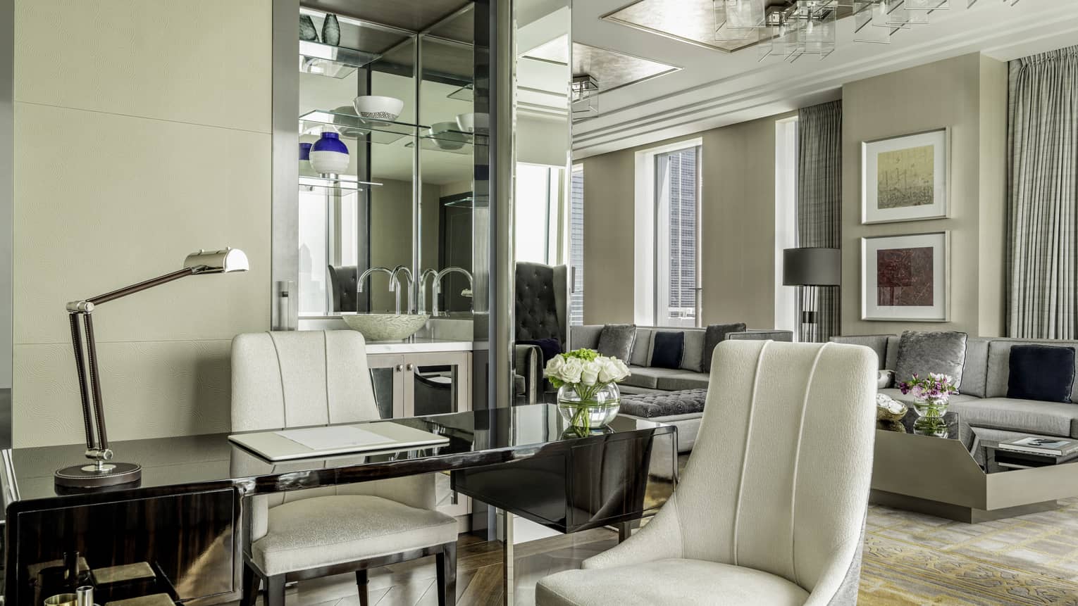 Elegant suite with a glass desk, plush white chairs, a modern lounge area with gray sofas, floral accents and decorative mirrors reflecting the chic decor