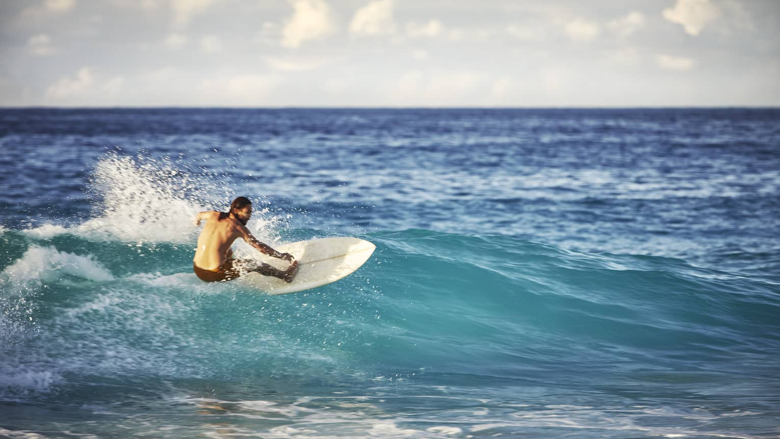 On an expanse of water, a surfer leans back on his board and surfs the face of a wave as white water splashes up behind him.