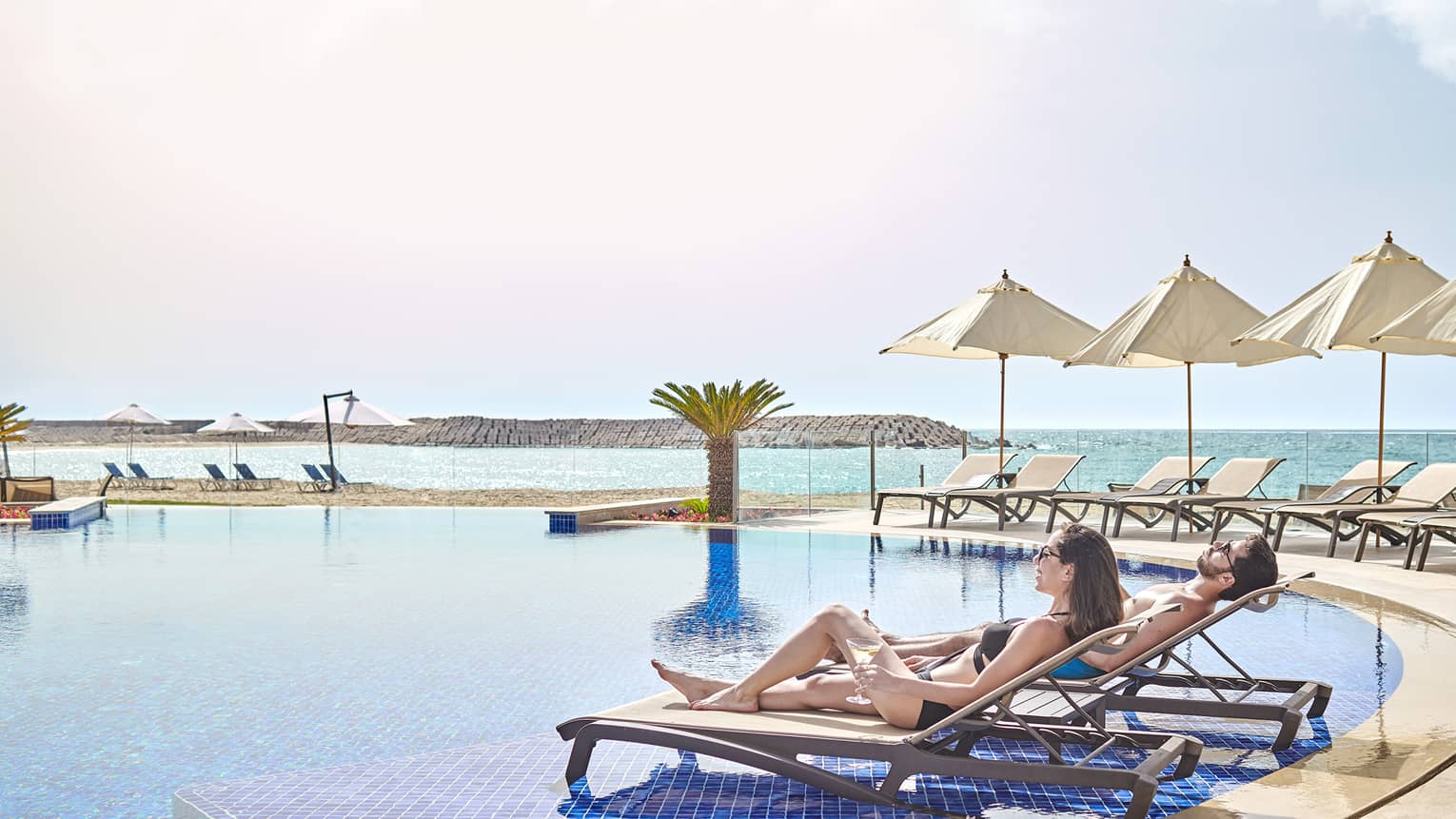A couple lounges by the pool with view of ocean, beach umbrellas 