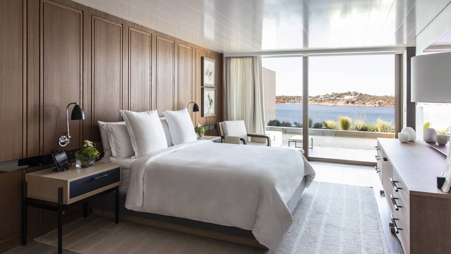 White king bed, wooden accent wall, contemporary night stand and dresser, wall of windows with sea view