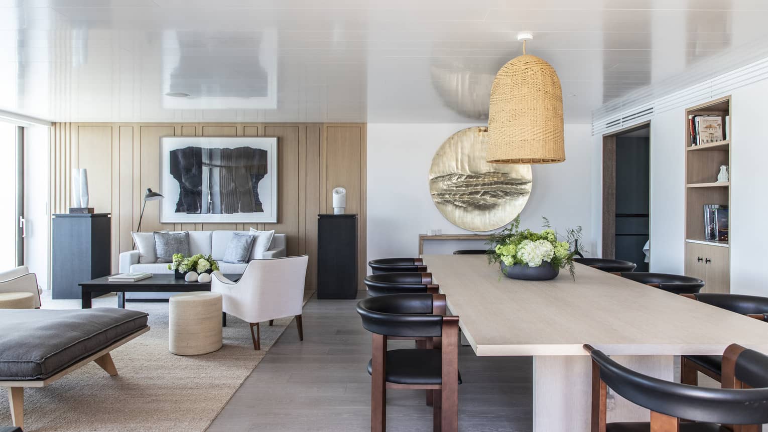 Light wood dining table, eight contemporary wood and black leather chairs, hanging wicker lamp, adjacent living room
