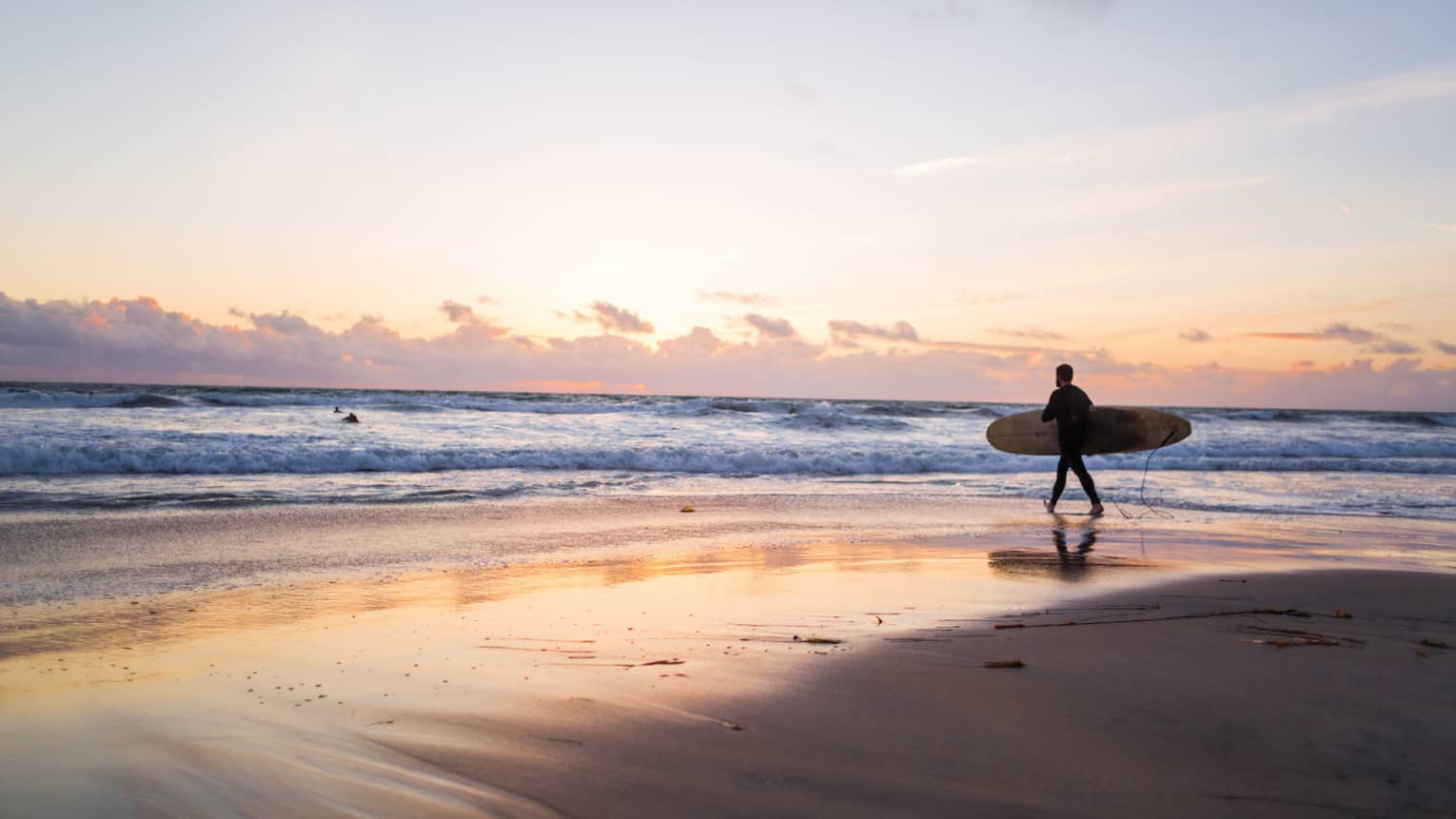 Under an expansive sky at sunset, clouds receding on the horizon, a surfer with a board heads into the glimmering ocean.