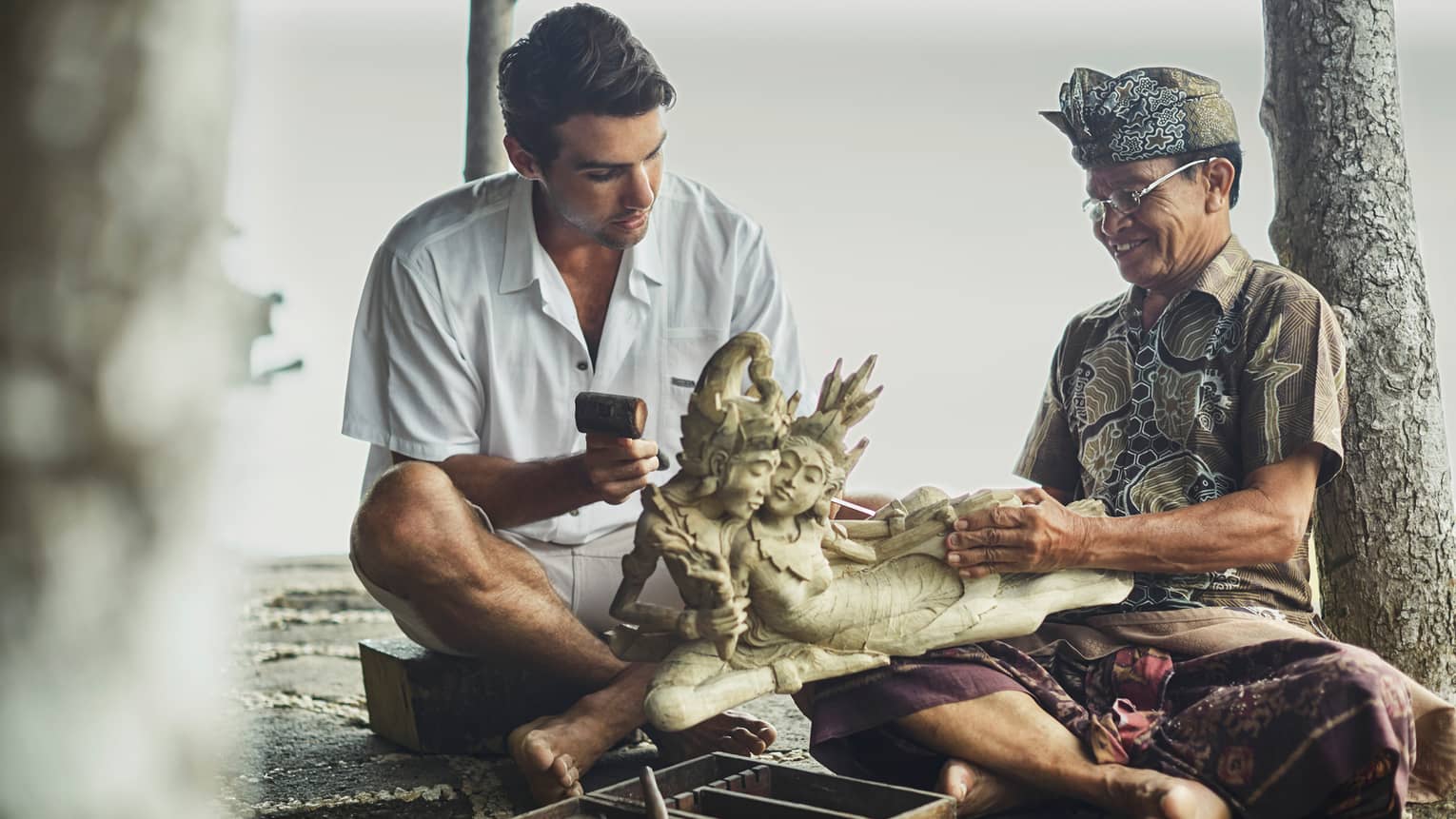 Man in white shirt sitting on mat with man in headdress and native costume holding carving