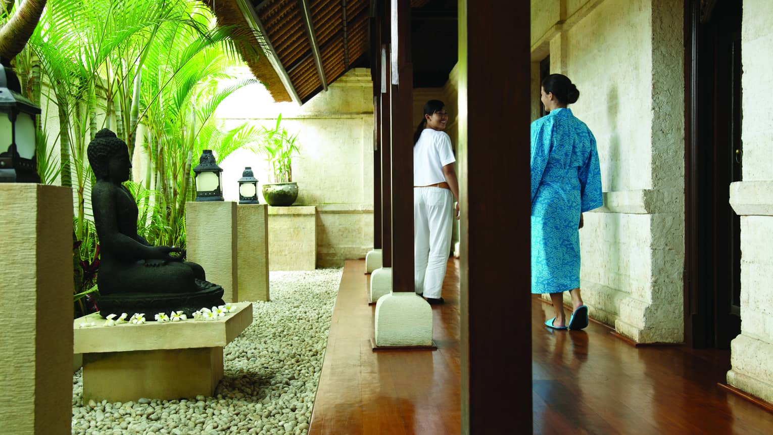Woman in blue robe walks down hallway past black Buddha statue, rock garden