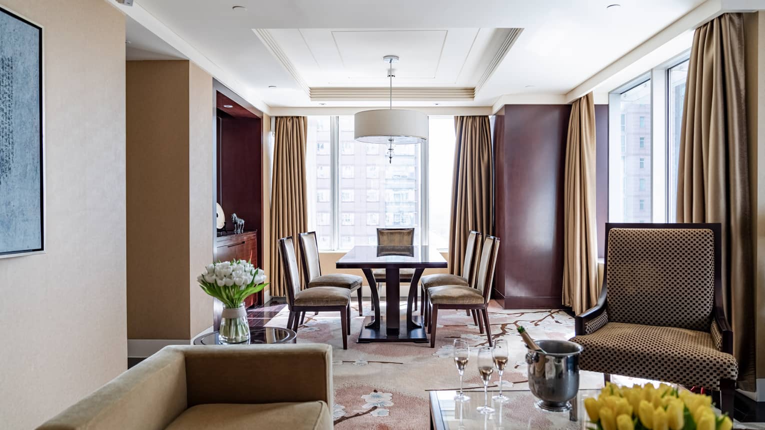 Tan couch and accent chair, glass coffee table, wooden dining table with four chairs, floor-to-ceiling windows