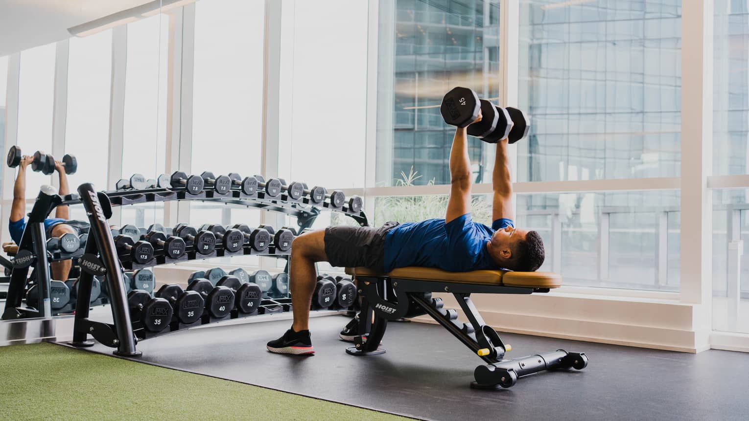 A man working out with free weights in a clean fitness room with large windows.