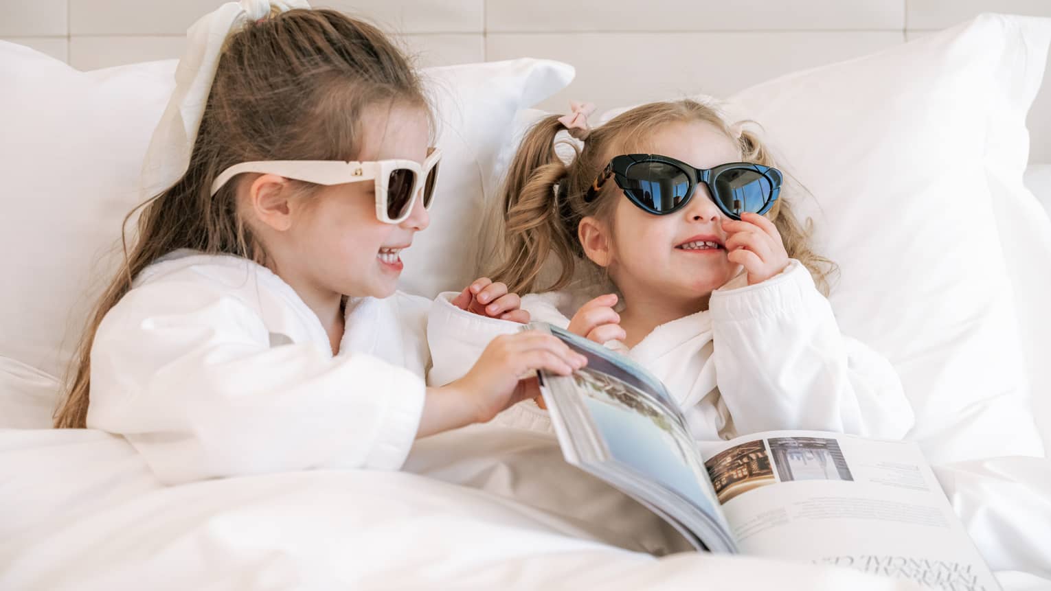Two young girls lounge in hotel bed wearing sunglasses
