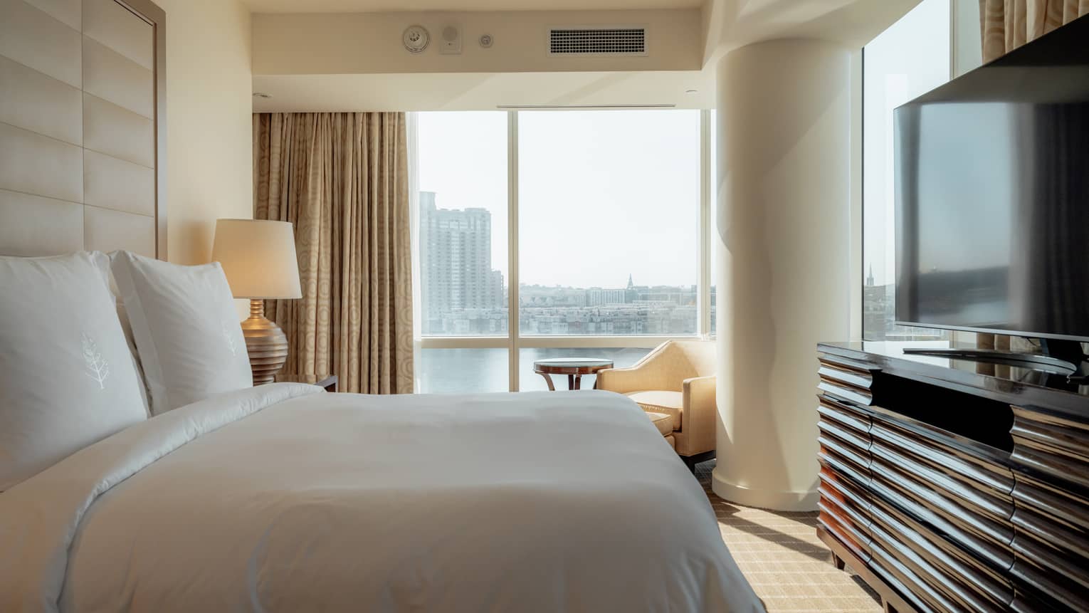Luxury hotel room with king bed, TV and dresser, and a chair and small table next to floor-to-ceiling window