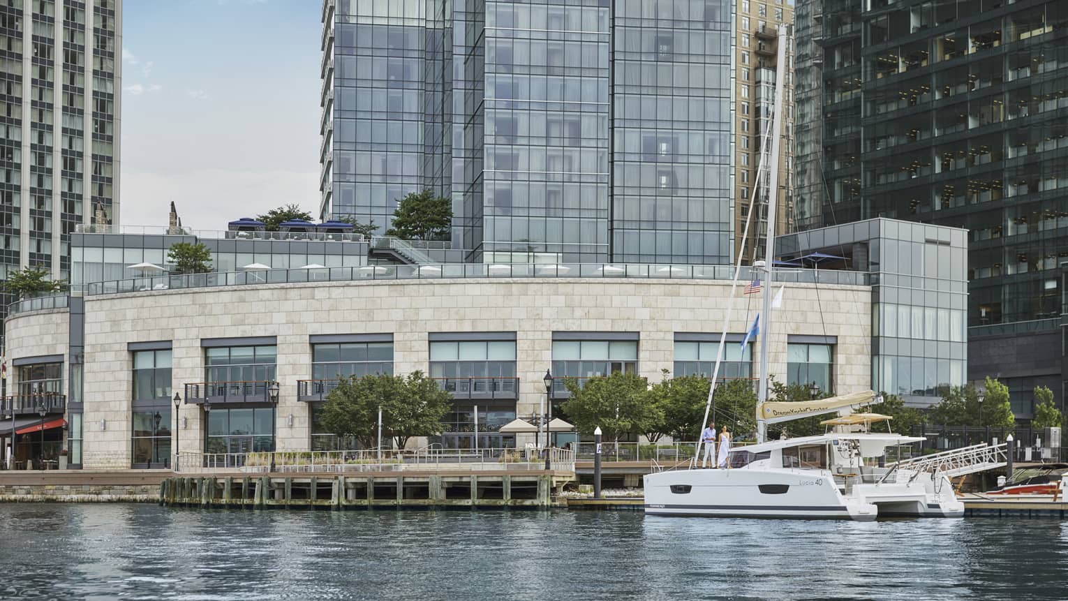 A sail boat on the water next to a large brick building and a pier, with tall skyscrapers in the background.
