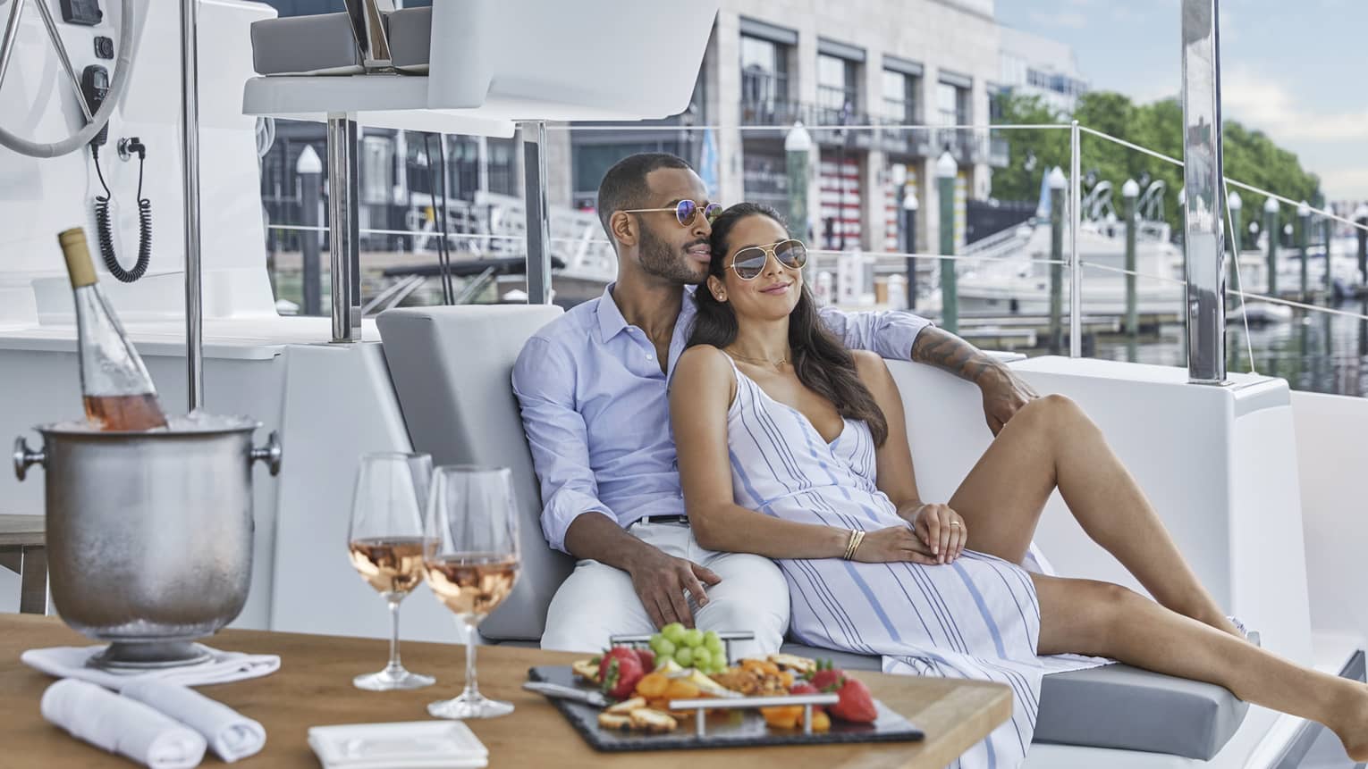 A man and a woman sitting closely together outside on a boat with wine, fresh fruit and bread on a table next to them.