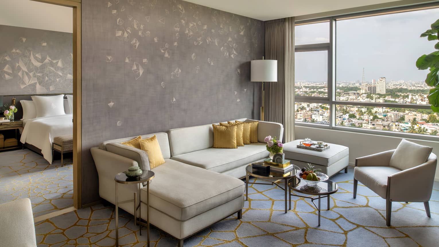 Guest room living area with cream-coloured sectional sofa and matching armchair, window with city views, doorway to bedroom