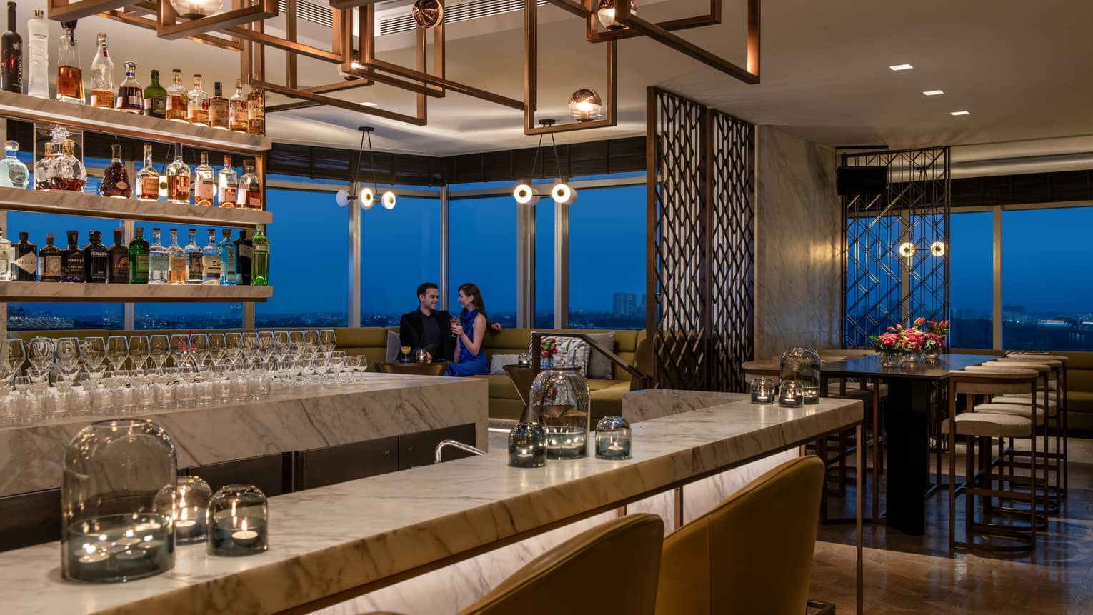 Two smiling people in formal wear sit in booth behind marble bar with liquor bottles on display