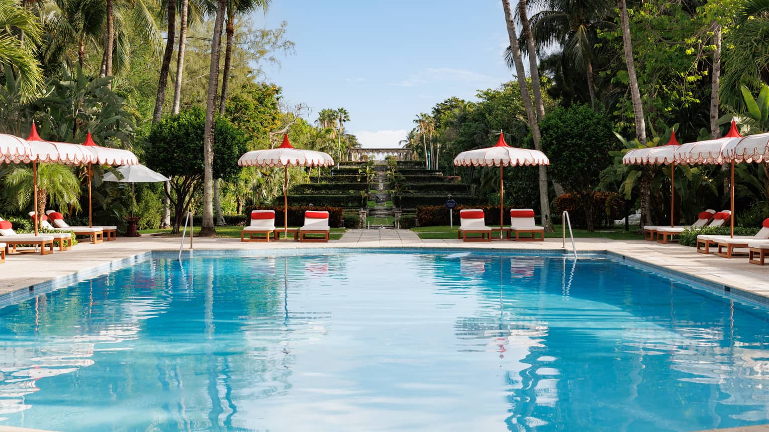 An outdoor pool at a tropical resort, flanked by white and red lounge chairs and umbrellas