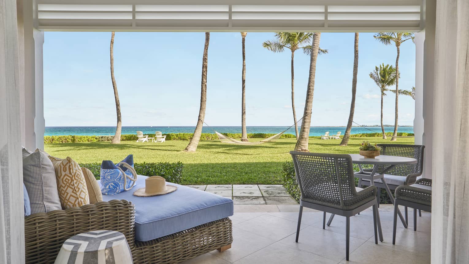 Outdoor patio with wicker lounge seating, dining table, and a view of a lawn with palm trees, hammock, and ocean