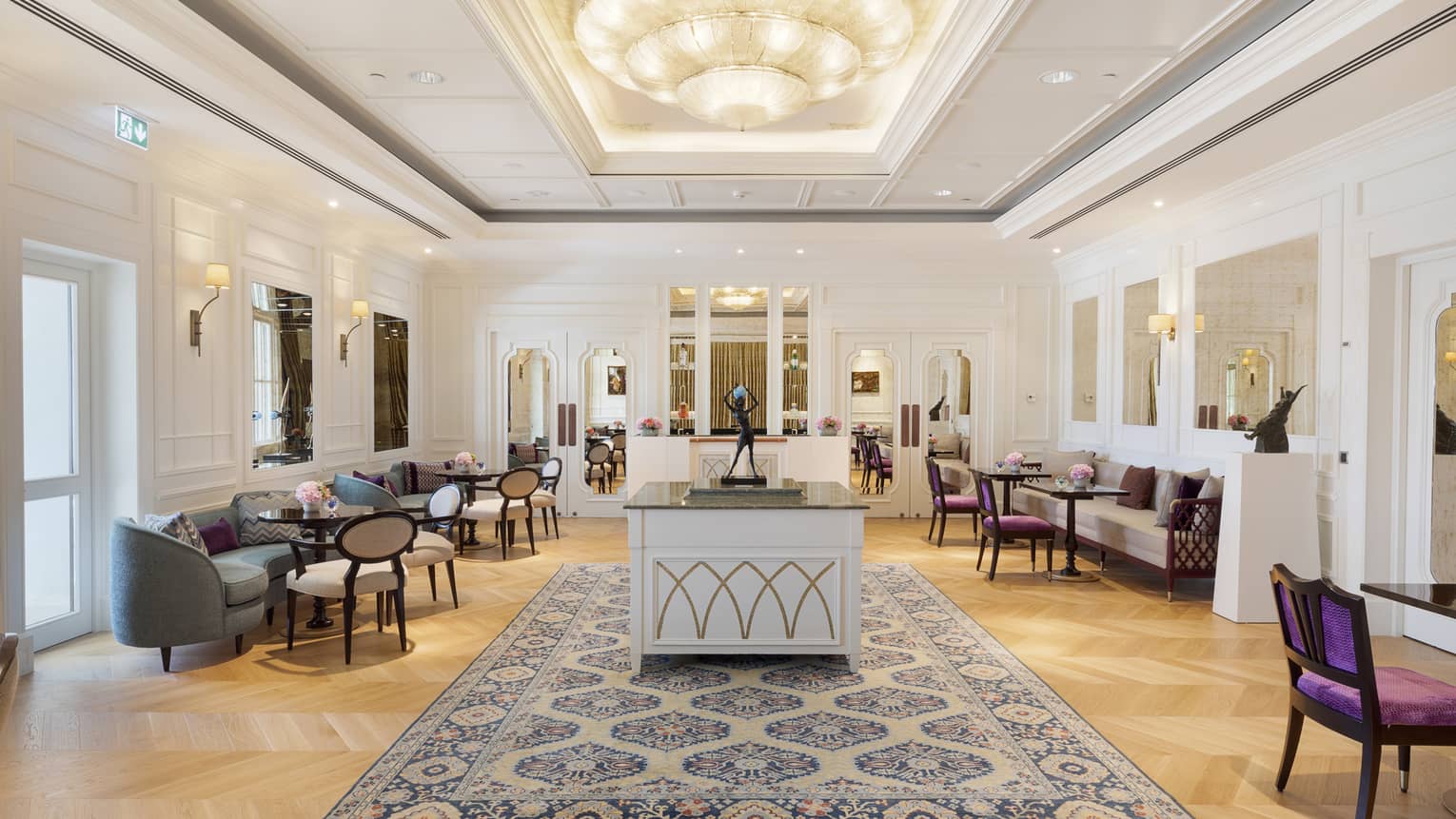 Dining room with artwork in the middle, chandelier overhead, walls lined with tables, sofas and wooden chairs