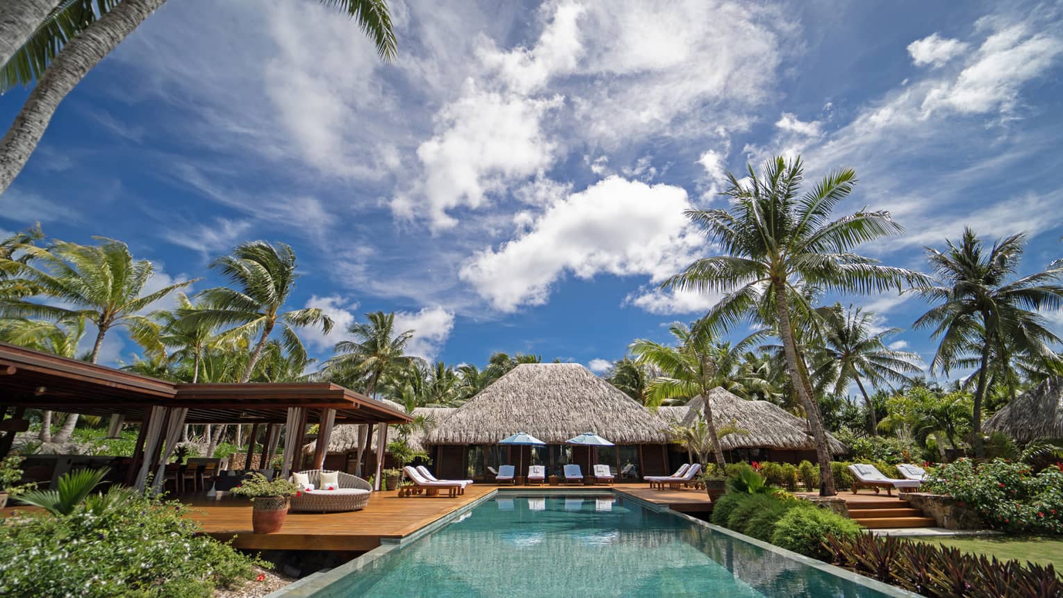 View from end of long rectangular pool, looking back across the pool to thatched-roof villa