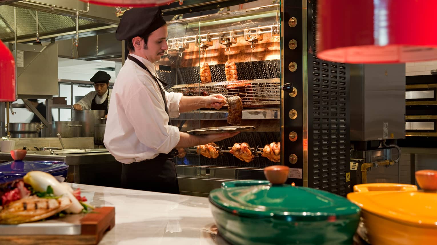 Cook takes meat out of large rotisserie in kitchen, green and yellow baking dishes with lids on counter