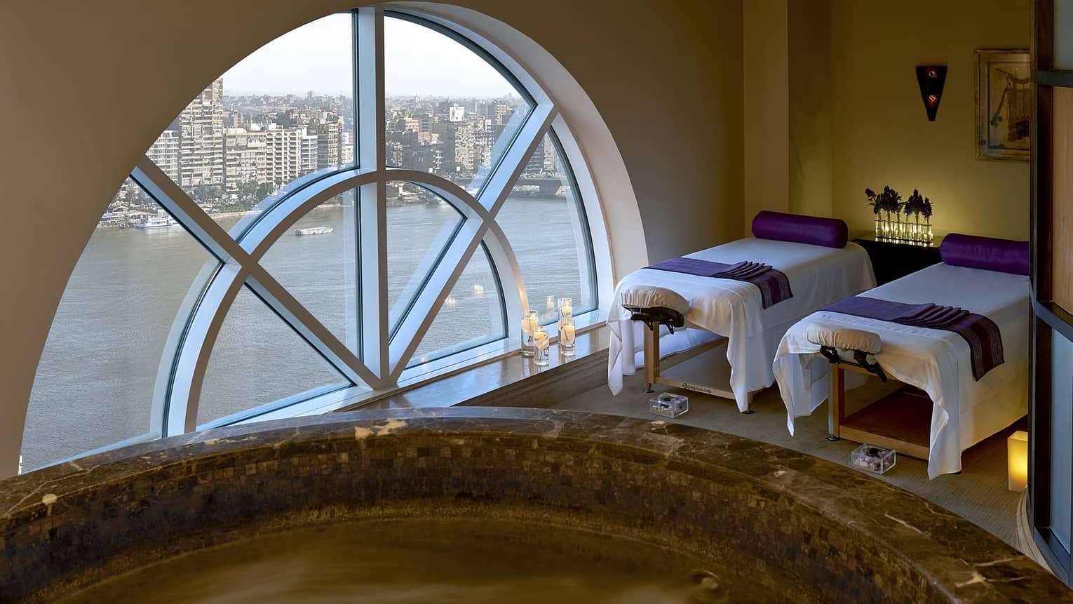 Two massage tables side-by-side in couples spa suite by lare crescent-shaped window overlooking Nile