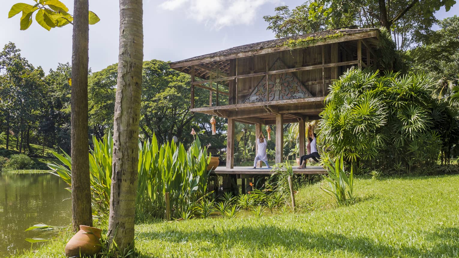 Yogi and two guests kneel with arms raised in yoga posed on pavilion deck near pond