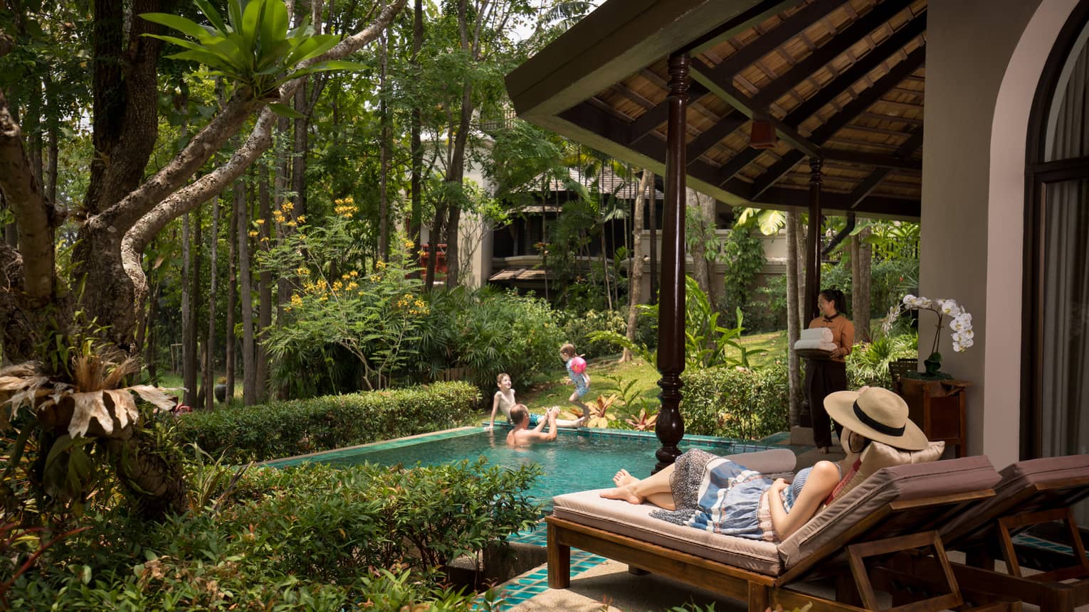 Person relaxing on a sun lounger beside a private pool while others play in the water and a staff member carries fresh towels nearby amidst lush greenery