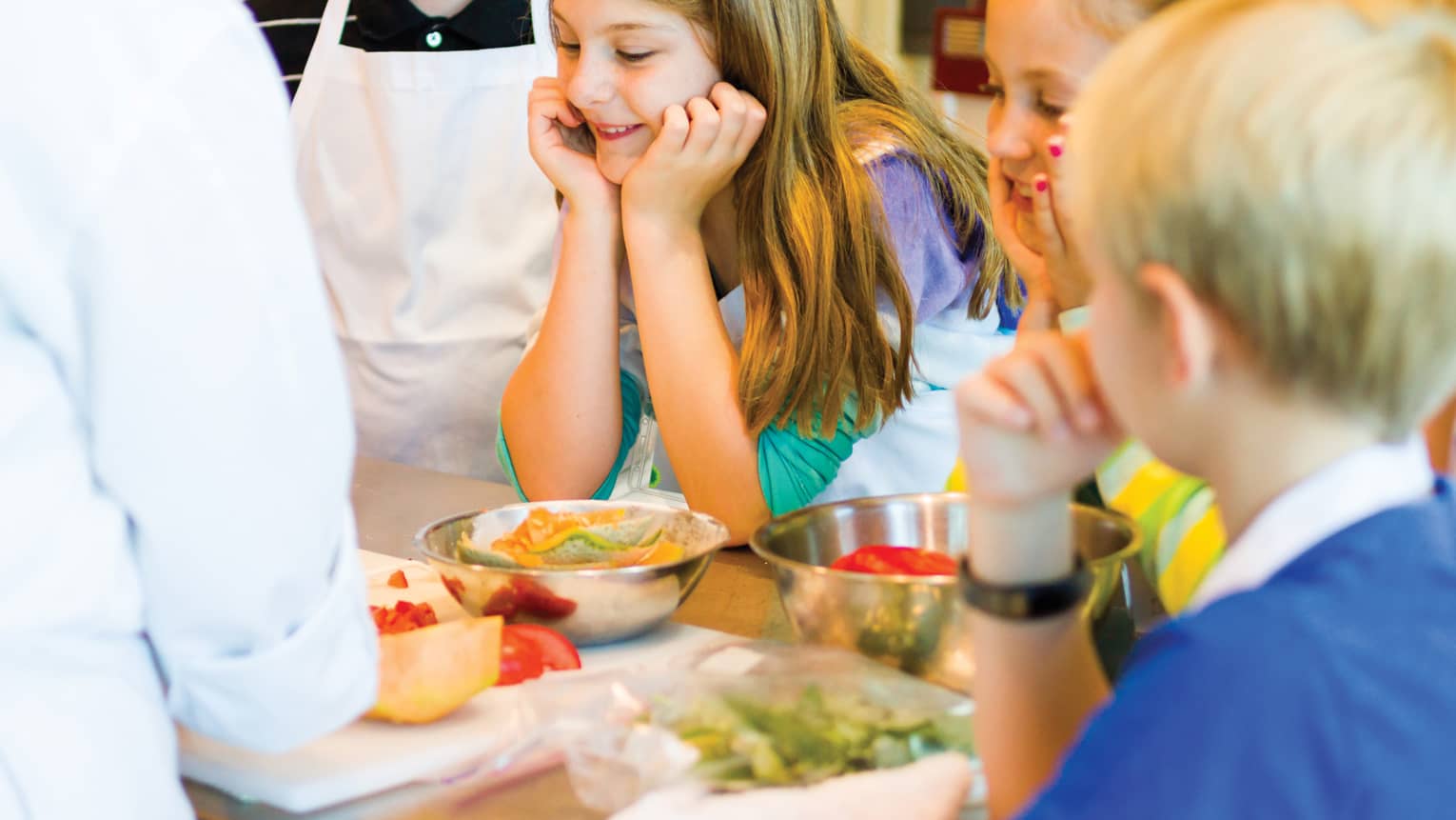 Smiling girl on apron rests chin on hands by other kids during Thai cooking class