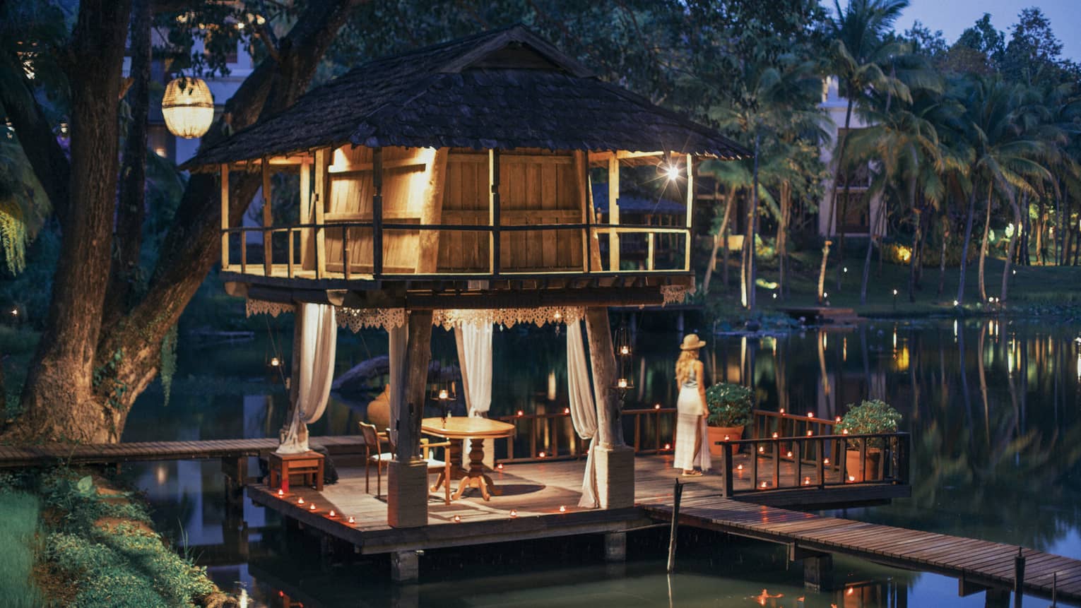 Person standing on a dock beside a candlelit lakeside pavilion with a thatched roof and draped curtains at dusk.