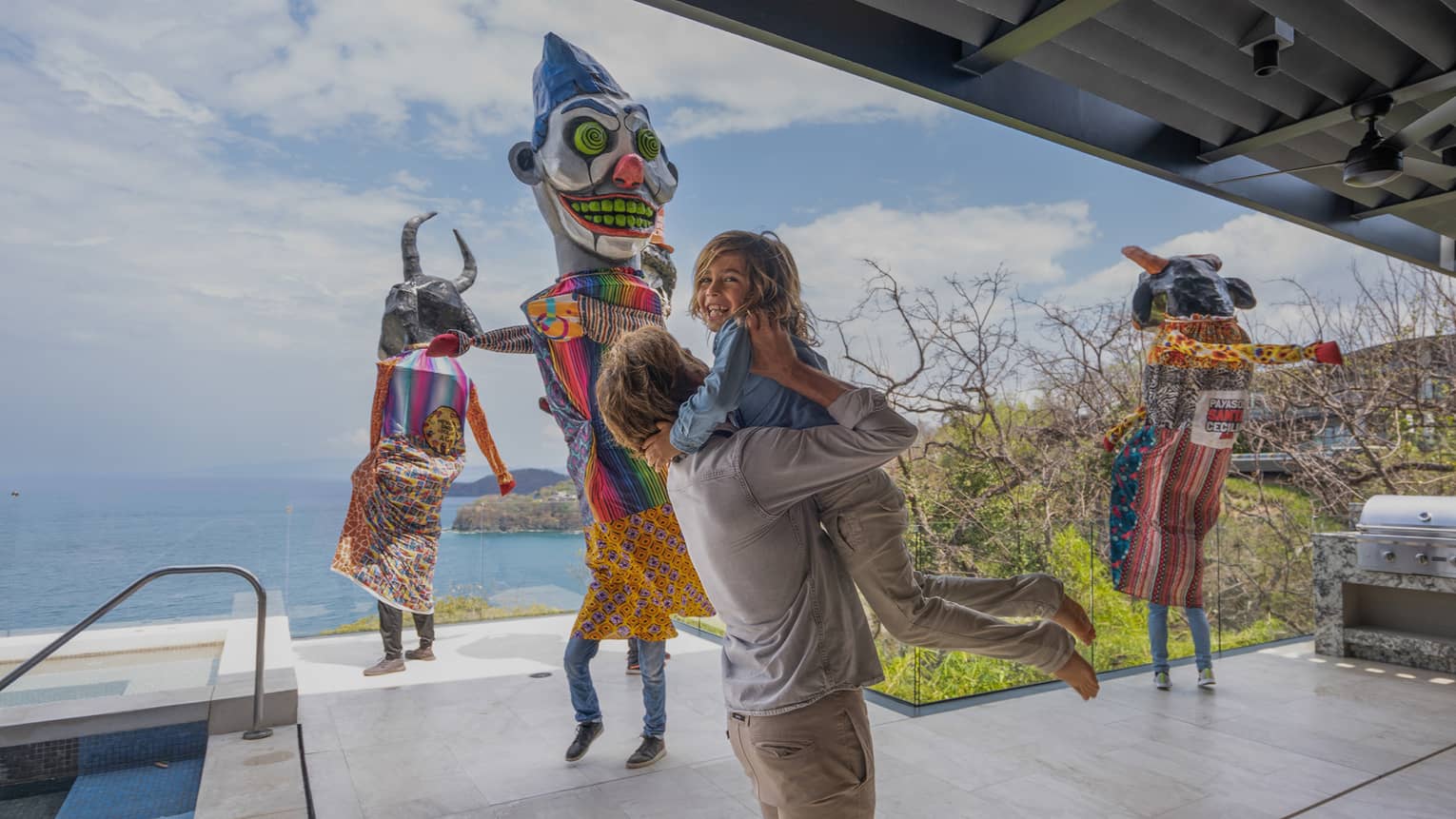 A young boy with blond curly hair is lifted up into the arms of his father as three people dressed in traditional Mexican costumes dance in the background