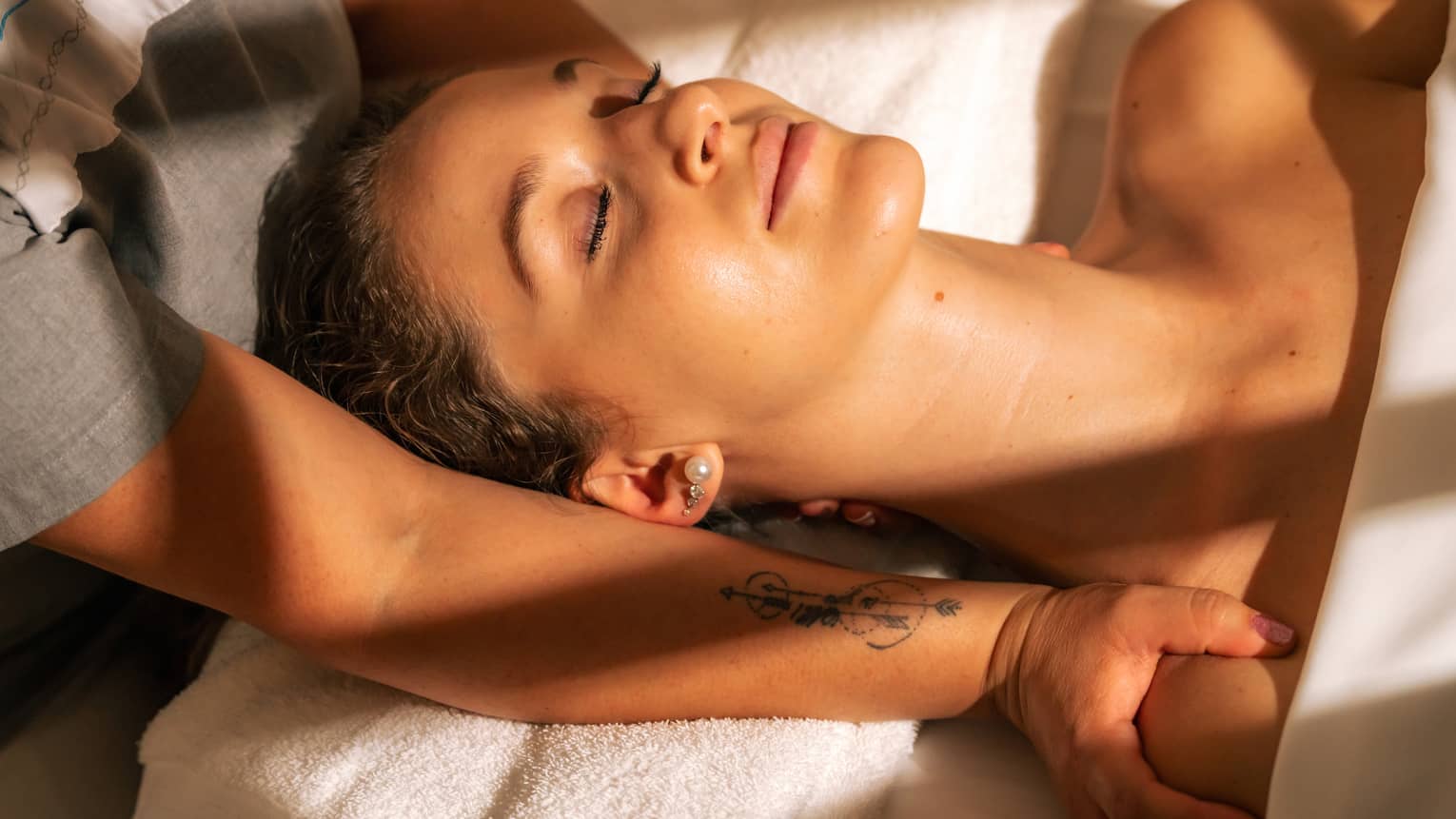 Person lies on their back covered from shoulders down by a white towel, their face lit by the sun, as a massage therapist massages their shoulders and neck