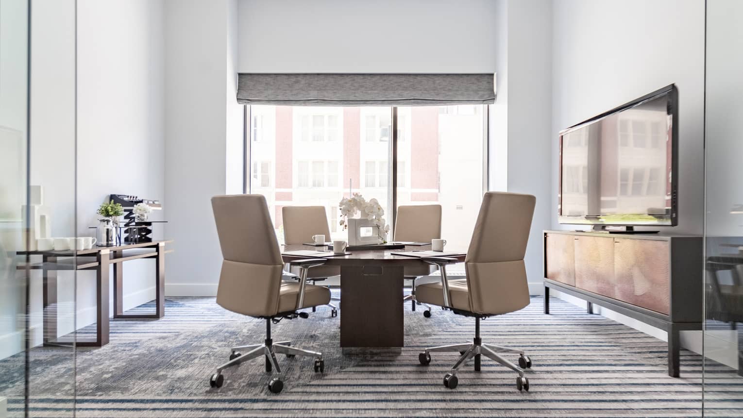 A well lit meeting room with a tv, a large window, a round table and four chairs, and a counter with cups and water.