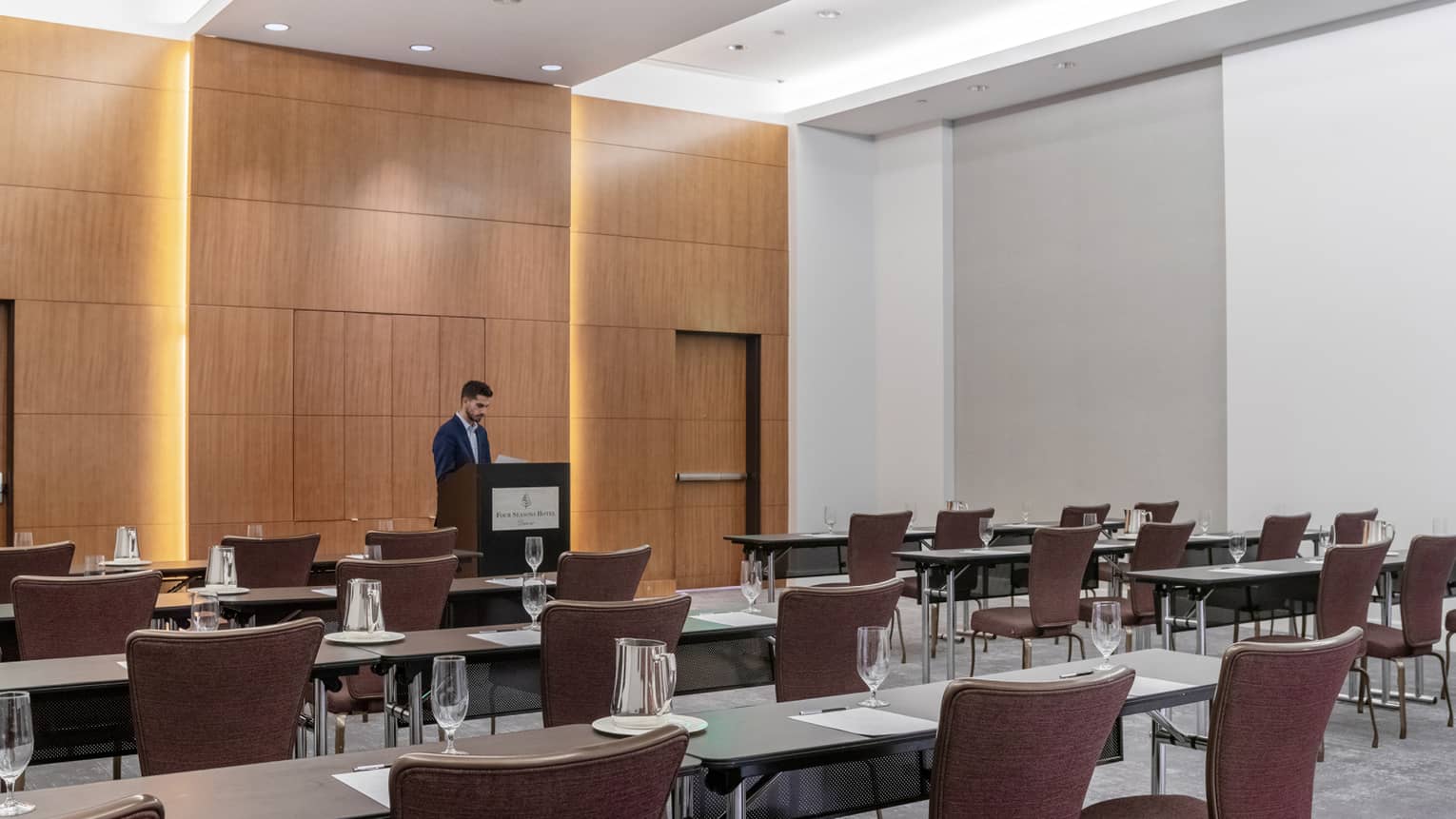 A man standing at a podium, rows of tables and brown chairs are in front of him.