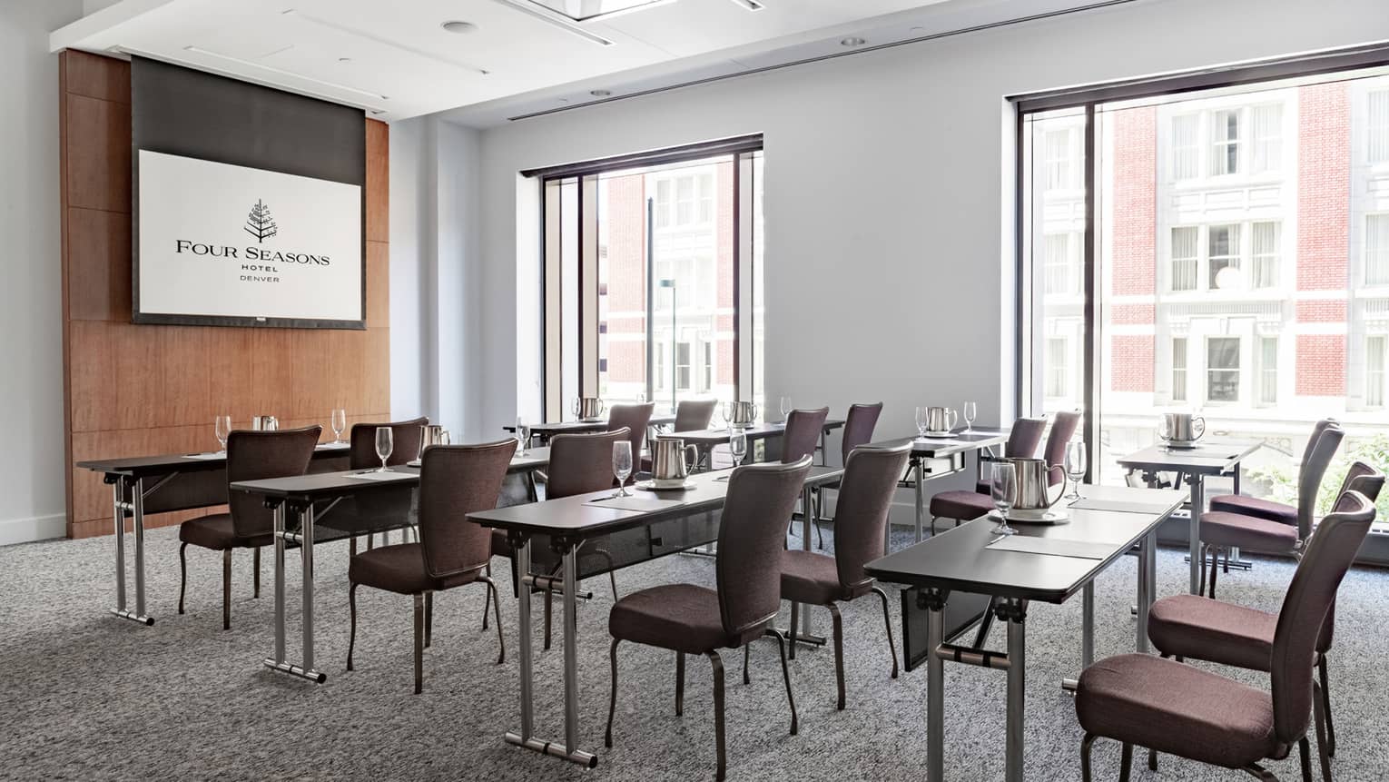 Five rows of desks and brown chairs facing a large TV on the wall, large windows are letting light into the room.