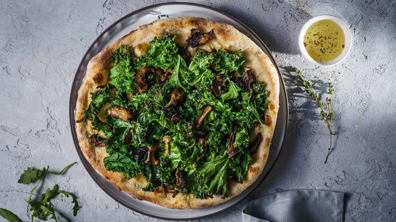 A flatbread pizza covered in greens and wild mushrooms, served on a white platter with garlic sauce.